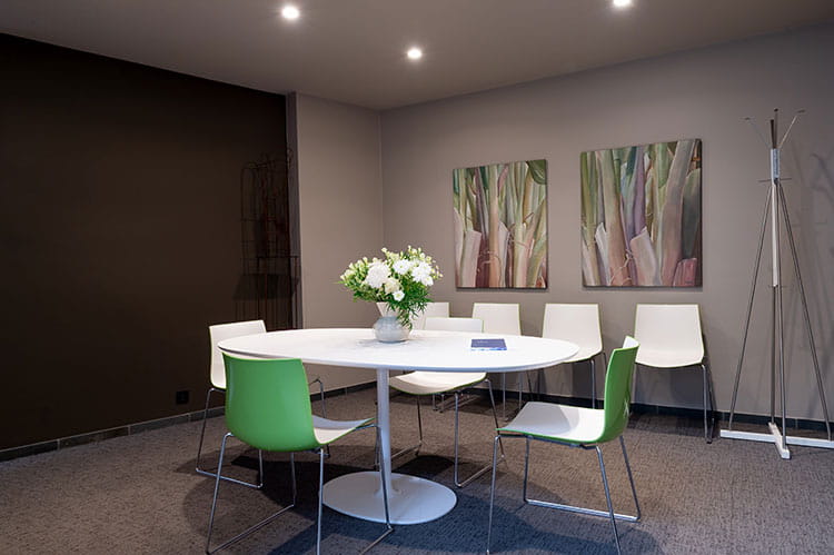 Interieur van een vergaderruimte met een ronde tafel en stoelen, een bloemenvaas op tafel, twee schilderijen aan de muur en een kapstok in de hoek.